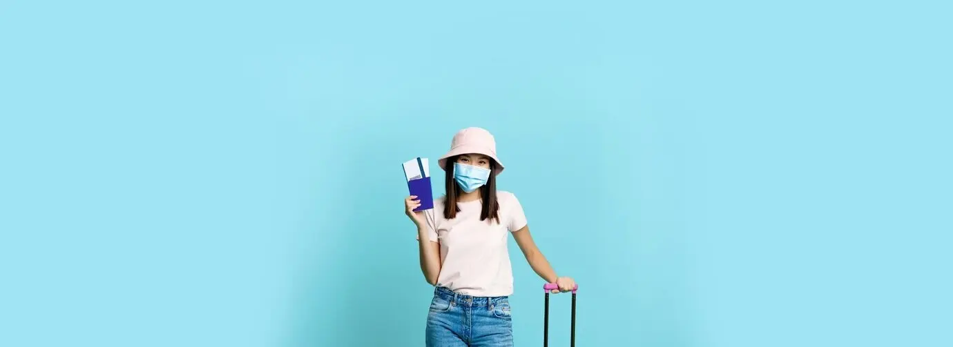Vertical shot of a cheerful Asian woman with a suitcase, wearing a face mask and displaying a passport and two tickets.